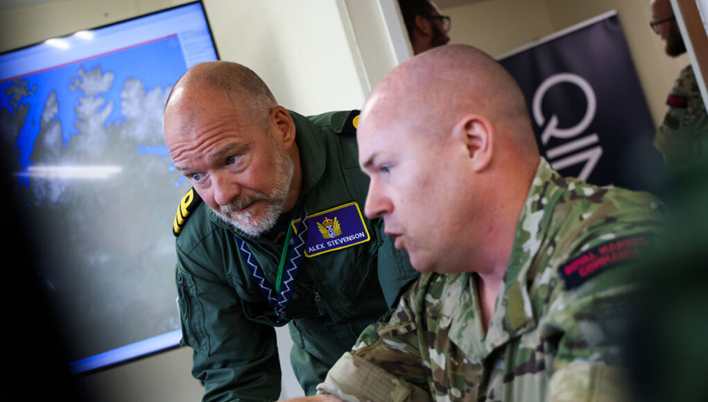 mcastMODimage Two soldiers lean over a desk, examining a large monitor showing a world map in a briefing room.