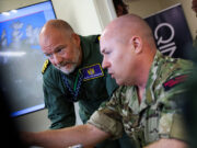 Inside Virtual Warrior – the Royal Navy’s secret war game Two soldiers lean over a desk, examining a large monitor showing a world map in a briefing room.