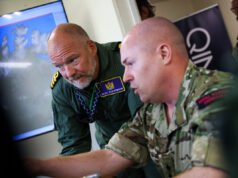 Two soldiers lean over a desk, examining a large monitor showing a world map in a briefing room.