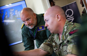 Inside Virtual Warrior – the Royal Navy’s secret war game Two soldiers lean over a desk, examining a large monitor showing a world map in a briefing room.