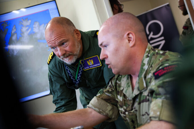 Two soldiers lean over a desk, examining a large monitor showing a world map in a briefing room.