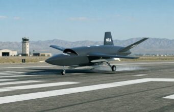 Unmanned military drone taxiing on a runway with a control tower and desert mountains in the background.