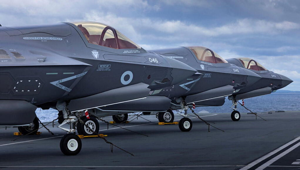 pwls Several gray military fighter jets parked in a row on a ship's flight deck, secured with chains with the ocean in the background.