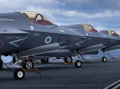 Several gray military fighter jets parked in a row on a ship's flight deck, secured with chains with the ocean in the background.