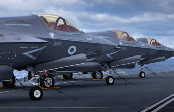 Several gray military fighter jets parked in a row on a ship's flight deck, secured with chains with the ocean in the background.