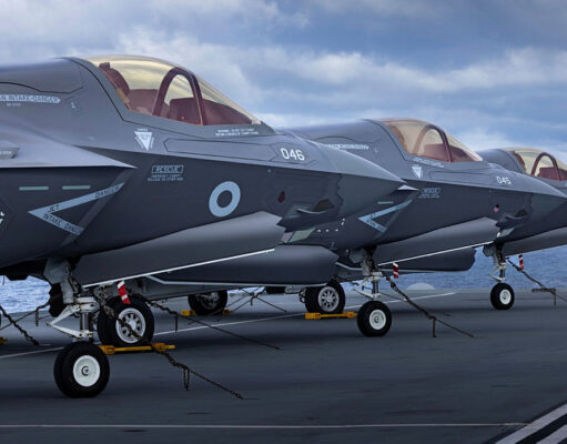 Several gray military fighter jets parked in a row on a ship's flight deck, secured with chains with the ocean in the background.