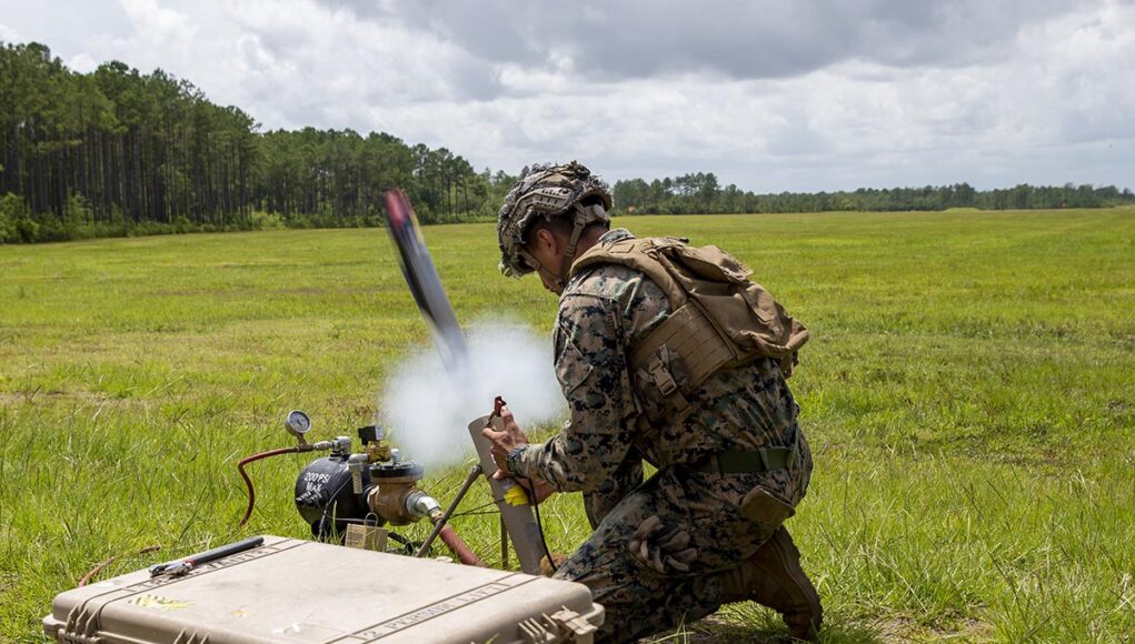 Soldier in camouflage kneels in a grassy field, handling a portable rocket launcher with a large rugged equipment case nearby.