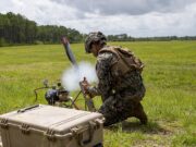 Soldier in camouflage kneels in a grassy field, handling a portable rocket launcher with a large rugged equipment case nearby.