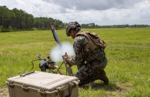 Soldier in camouflage kneels in a grassy field, handling a portable rocket launcher with a large rugged equipment case nearby.