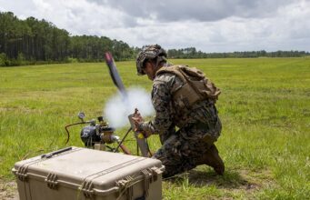 US approves Switchblade drone sale to Greece Soldier in camouflage kneels in a grassy field, handling a portable rocket launcher with a large rugged equipment case nearby.