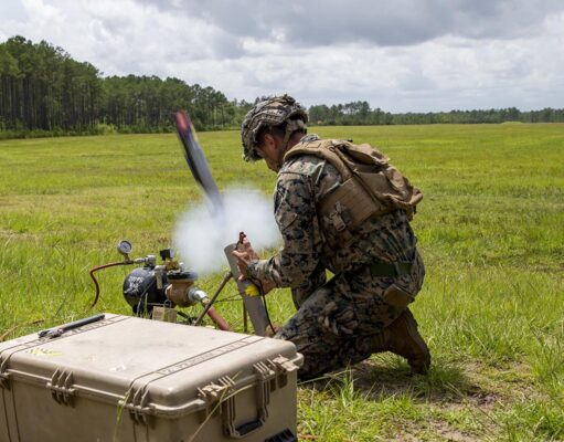 Soldier in camouflage kneels in a grassy field, handling a portable rocket launcher with a large rugged equipment case nearby.