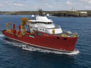 UK’s Trinity House awards huge ship deal to Norway’s VARD Red offshore support vessel with white superstructure and a yellow crane array, moving through open water; helicopter on the forward helipad and a lighthouse visible on a rocky coastline in the background, vessel marked VARD 9 601.