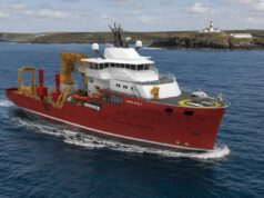 UK’s Trinity House awards huge ship deal to Norway’s VARD Red offshore support vessel with white superstructure and a yellow crane array, moving through open water; helicopter on the forward helipad and a lighthouse visible on a rocky coastline in the background, vessel marked VARD 9 601.