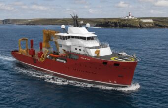 UK’s Trinity House awards huge ship deal to Norway’s VARD Red offshore support vessel with white superstructure and a yellow crane array, moving through open water; helicopter on the forward helipad and a lighthouse visible on a rocky coastline in the background, vessel marked VARD 9 601.