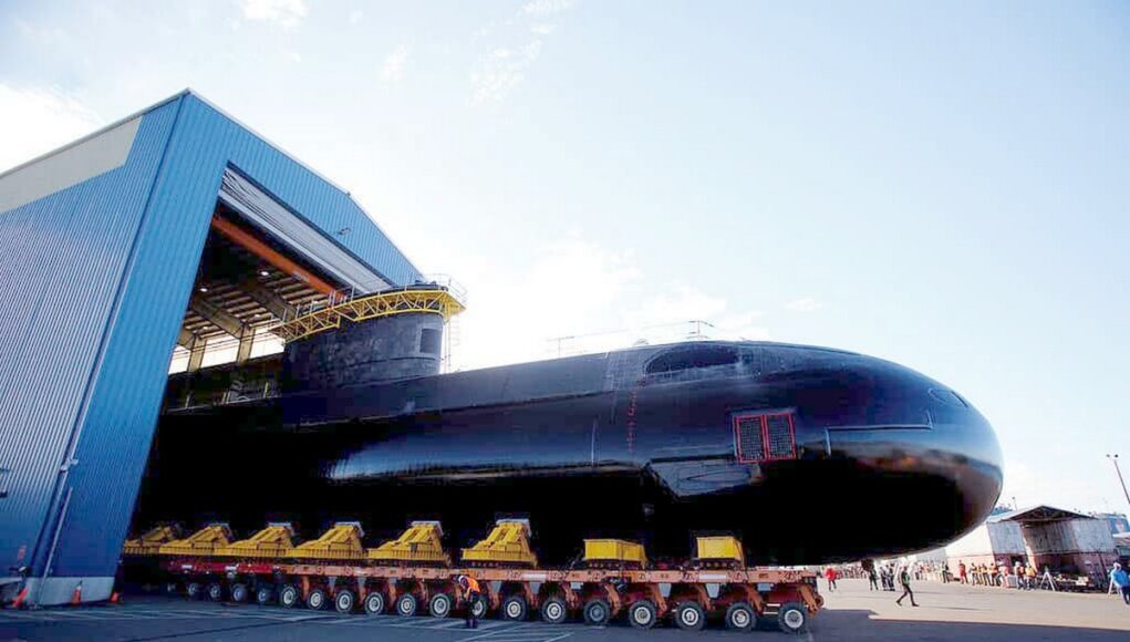 vic1 Large black submarine on a long, multi-wheeled transport trailer outside a blue hangar, with workers nearby.
