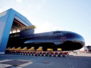 Babcock wins six-year extension for Canada sub support Large black submarine on a long, multi-wheeled transport trailer outside a blue hangar, with workers nearby.