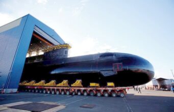 Babcock wins six-year extension for Canada sub support Large black submarine on a long, multi-wheeled transport trailer outside a blue hangar, with workers nearby.