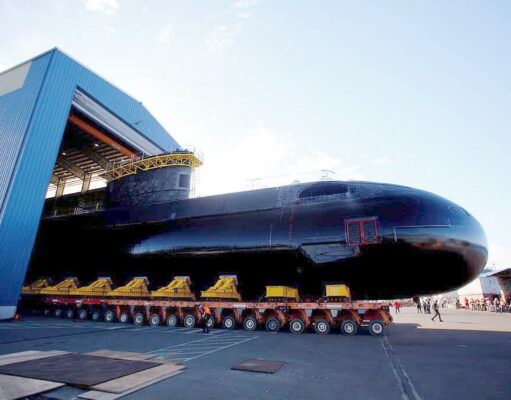 Babcock wins six-year extension for Canada sub support Large black submarine on a long, multi-wheeled transport trailer outside a blue hangar, with workers nearby.