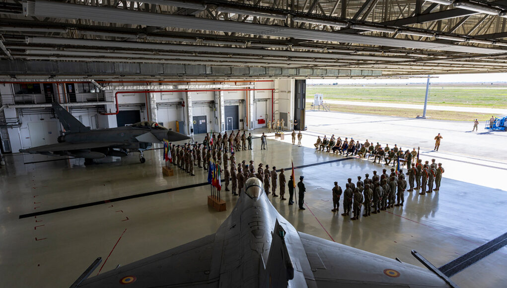 121_EAW20260731-0110-HOTO_Ceremony_-0699 Inside a large military hangar, a fighter jet sits on the floor while soldiers stand in formation around flags for a ceremony.