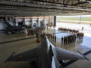 Inside a large military hangar, a fighter jet sits on the floor while soldiers stand in formation around flags for a ceremony.