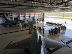 RAF hands Romania air policing mission to Spain Inside a large military hangar, a fighter jet sits on the floor while soldiers stand in formation around flags for a ceremony.