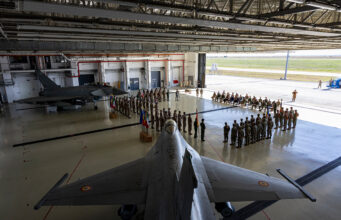 RAF hands Romania air policing mission to Spain Inside a large military hangar, a fighter jet sits on the floor while soldiers stand in formation around flags for a ceremony.