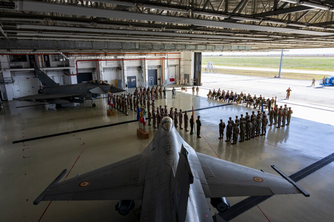 RAF hands Romania air policing mission to Spain Inside a large military hangar, a fighter jet sits on the floor while soldiers stand in formation around flags for a ceremony.