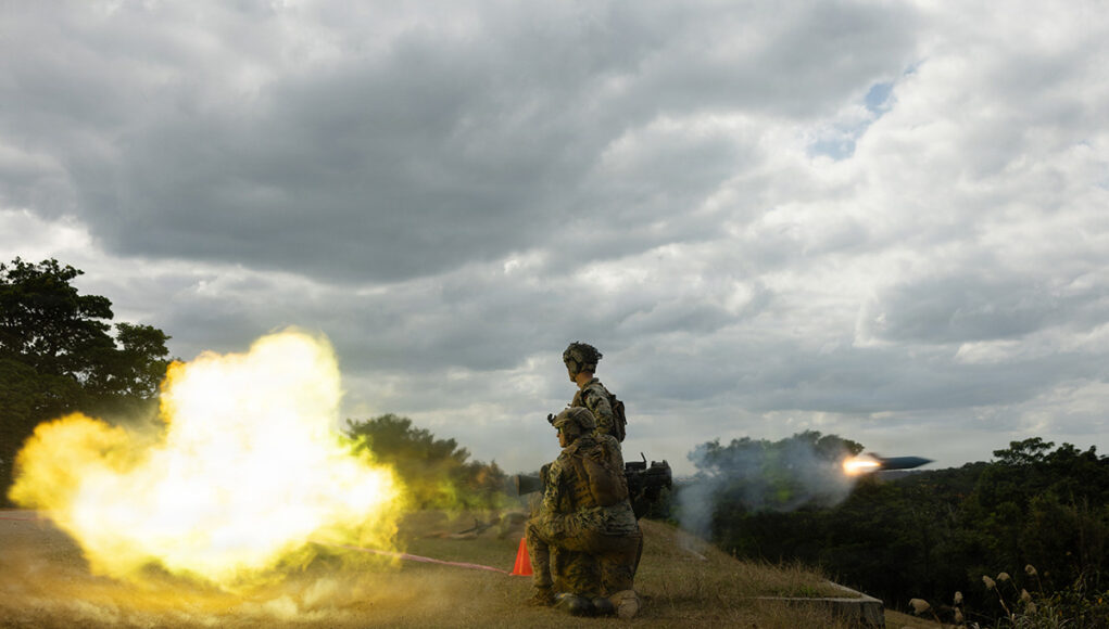 Two soldiers in camouflage kneeling on a grassy hill as a rocket launcher fires, bright yellow flame and smoke against a cloudy sky.