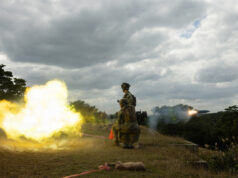 Two soldiers in camouflage kneeling on a grassy hill as a rocket launcher fires, bright yellow flame and smoke against a cloudy sky.