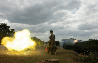 U.S. Marines open fire in first overhead drill on Okinawa Two soldiers in camouflage kneeling on a grassy hill as a rocket launcher fires, bright yellow flame and smoke against a cloudy sky.