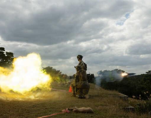 U.S. Marines open fire in first overhead drill on Okinawa Two soldiers in camouflage kneeling on a grassy hill as a rocket launcher fires, bright yellow flame and smoke against a cloudy sky.