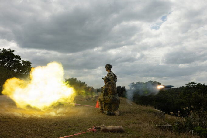 U.S. Marines open fire in first overhead drill on Okinawa Two soldiers in camouflage kneeling on a grassy hill as a rocket launcher fires, bright yellow flame and smoke against a cloudy sky.