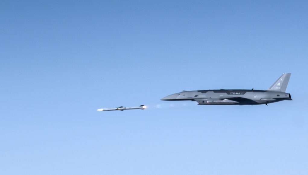 Large gray military aircraft flying high with a small white drone beside it in clear blue sky.