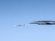 US Air Force drone fires AIM-120 in live test Large gray military aircraft flying high with a small white drone beside it in clear blue sky.