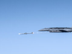 US Air Force drone fires AIM-120 in live test Large gray military aircraft flying high with a small white drone beside it in clear blue sky.