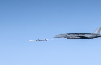 US Air Force drone fires AIM-120 in live test Large gray military aircraft flying high with a small white drone beside it in clear blue sky.