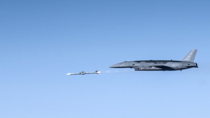 US Air Force drone fires AIM-120 in live test Large gray military aircraft flying high with a small white drone beside it in clear blue sky.