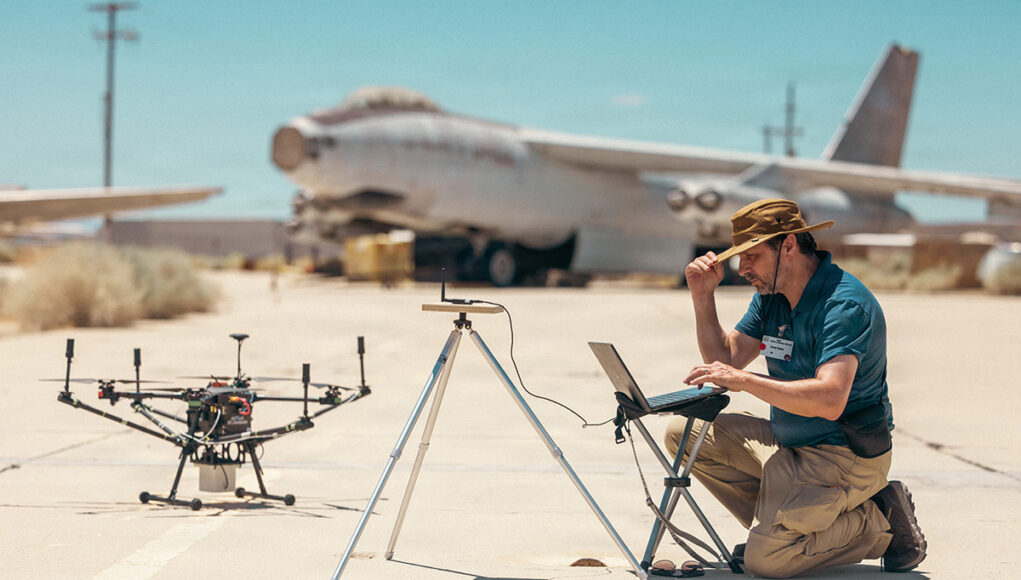 Man kneels on a sunlit tarmac, using a laptop beside a on-ground drone and a tripod setup, with a vintage aircraft in the desert background.