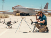 Drones printed and flown in a day at US test base Man kneels on a sunlit tarmac, using a laptop beside a on-ground drone and a tripod setup, with a vintage aircraft in the desert background.