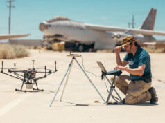 Drones printed and flown in a day at US test base Man kneels on a sunlit tarmac, using a laptop beside a on-ground drone and a tripod setup, with a vintage aircraft in the desert background.