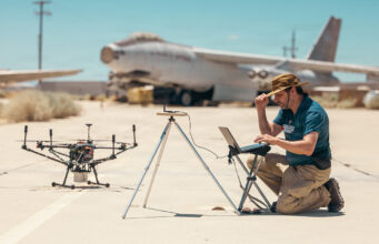 Drones printed and flown in a day at US test base Man kneels on a sunlit tarmac, using a laptop beside a on-ground drone and a tripod setup, with a vintage aircraft in the desert background.