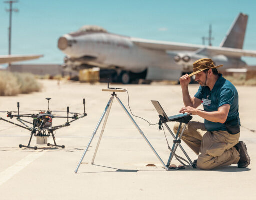 Drones printed and flown in a day at US test base Man kneels on a sunlit tarmac, using a laptop beside a on-ground drone and a tripod setup, with a vintage aircraft in the desert background.