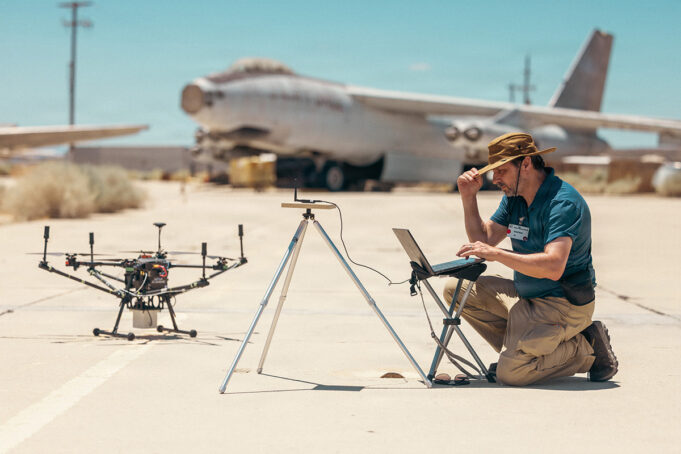 Man kneels on a sunlit tarmac, using a laptop beside a on-ground drone and a tripod setup, with a vintage aircraft in the desert background.