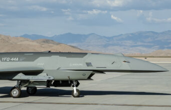 New American combat drones armed and flown in desert Close-up side view of a gray military jet on a taxiway, with arid mountains in the distance under a blue sky with clouds.