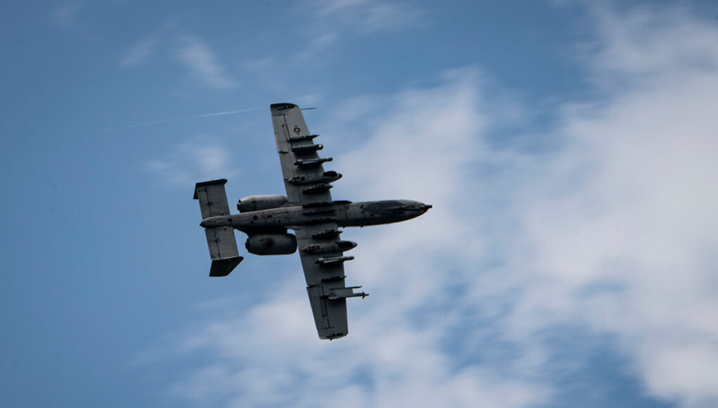 260729-F-XU250-1218 Underside view of a gray four-engine propeller aircraft (C-130) flying overhead against a blue sky.