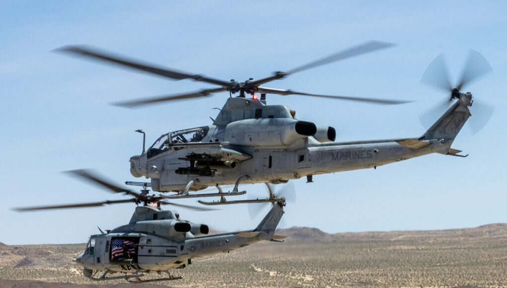 690325-H-1_Pt2_DVIDS-Photo-7768567-61674b-original-1784751234 Two US Marine Corps helicopters hovering low over a desert landscape with blurred rotor blades and a visible American flag decal on one side.