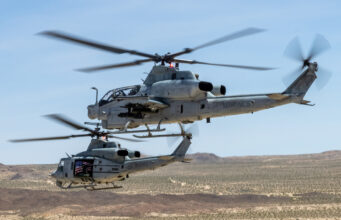 US Marine Corps gunships to get Link 16 upgrade Two US Marine Corps helicopters hovering low over a desert landscape with blurred rotor blades and a visible American flag decal on one side.