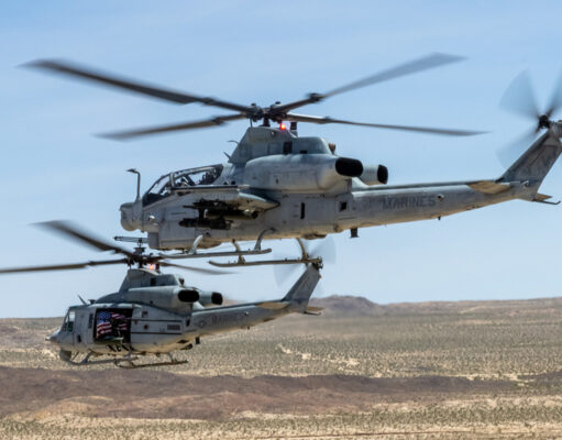 US Marine Corps gunships to get Link 16 upgrade Two US Marine Corps helicopters hovering low over a desert landscape with blurred rotor blades and a visible American flag decal on one side.