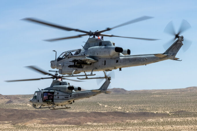 US Marine Corps gunships to get Link 16 upgrade Two US Marine Corps helicopters hovering low over a desert landscape with blurred rotor blades and a visible American flag decal on one side.