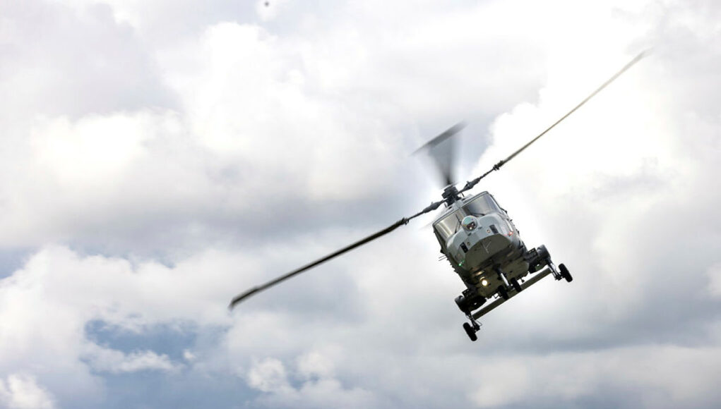 Gray military helicopter in flight above treeline under a cloudy sky, rotor blades blurred.
